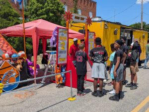 A group of kids wait in line at a street fair challenge, with a "Hang Tuff" game booth, stuffed prizes, and a yellow food truck in the background.