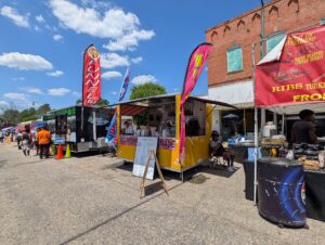 Food trucks and vendor stalls lined up on a sunny street, with people ordering food and colorful banners advertising items like lemonade, ribs, and funnel cakes.