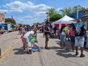 People gather at a street festival with food trucks, vendor tents, and families enjoying a sunny day. Some are in line, others walk or care for children in strollers.