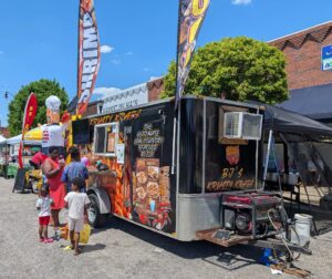 A family stands in front of a food truck named BJ’s Krusty Krabs, with seafood menu items and colorful banners, at an outdoor market on a sunny day.
