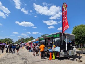 People gather at an outdoor food festival with colorful food trucks and a prominent funnel cake stand under a bright blue, partly cloudy sky.