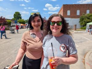 Two women smiling outdoors at a street fair on a sunny day, one holding a drink, both wearing sunglasses, with people and booths in the background.