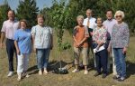 A group of people standing outdoors around a young tree, with greenery and trees in the background on a sunny day.
