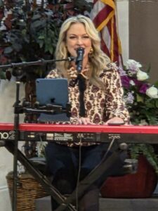A woman with long blonde hair plays a keyboard and sings into a microphone, with flowers and an American flag in the background.
