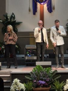 Three people holding microphones perform on a church stage with plants in front, audio equipment behind, and a large cross draped with purple cloth in the background.
