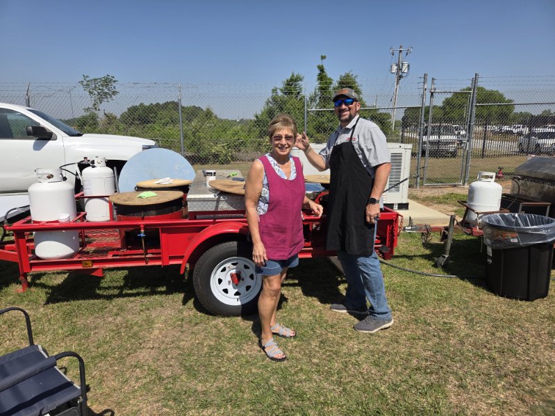 Two people wearing aprons stand next to a red trailer with cooking equipment outdoors on a sunny day.