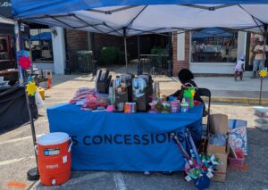 Outdoor concession stand under blue canopy, featuring drink machines, cups, toys, snacks, and a large orange cooler. Person sits behind the table; street and buildings in background.