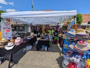 Outdoor market stall with hats, keychains, Greek life apparel, sports jerseys, accessories, and decorative items displayed under a white canopy. A person stands in the middle under the tent.