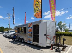 A food truck with colorful flags advertises hot dogs, corn dogs, sausage dogs, chili cheese fries, and lemonade. A person sits nearby under a sunny sky.