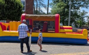 A man and a boy stand near a colorful inflatable bounce house on a sunny day, with trees and buildings in the background.