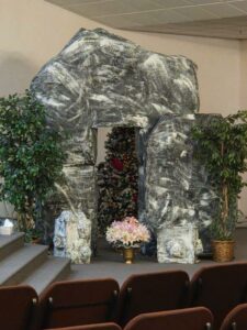 Large fake stone archway with plants, flowers, and a decorated Christmas tree visible through the opening, set up indoors near rows of brown chairs.