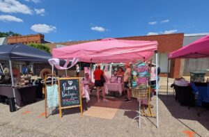 Outdoor market scene with people under a pink tent labeled "Rumor Boutique," displaying clothing and accessories, on a sunny day with a blue sky and brick buildings in the background.