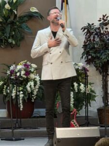 A man in a light blazer sings into a microphone, standing on a stage with plants and flowers in the background.