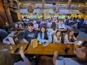 Group of people sitting at a wooden table in a busy, rustic restaurant with multiple diners in the background.