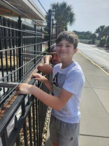 Smiling boy in a white shirt stands by a black metal fence on a sunny sidewalk, with another person behind him and palm trees in the background.