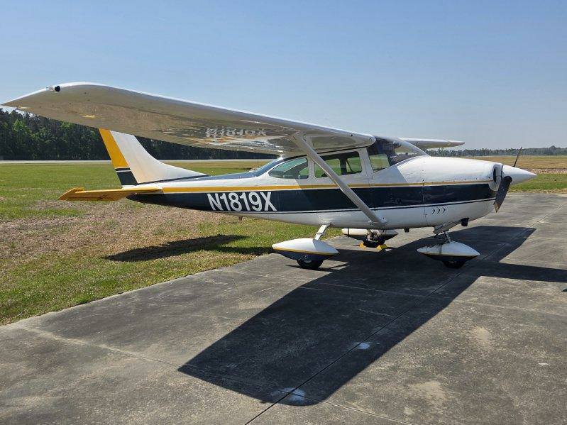 Small single-engine airplane parked on a tarmac under a clear blue sky, with grass fields in the background.