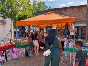 People browsing an outdoor market stall with toys, stuffed animals, handbags, and backpacks displayed under an orange tent on a sunny day.