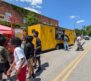 People line up at a yellow food truck and nearby carnival booth on a sunny street, with children and adults enjoying the outdoor event.