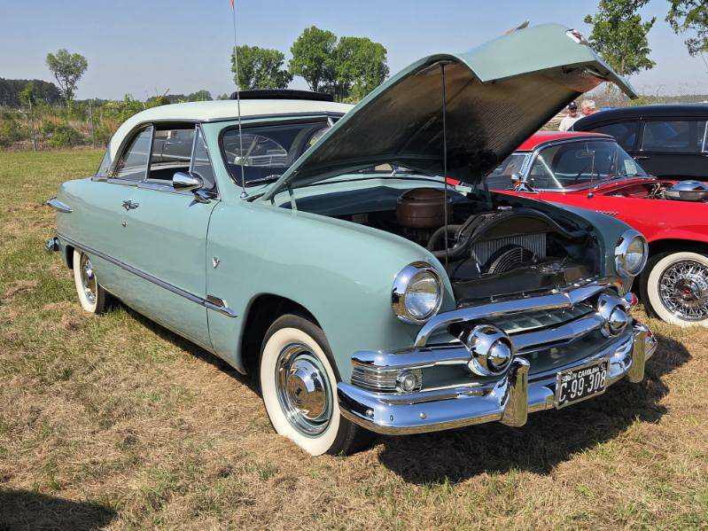 A vintage light blue car with its hood open, parked on grass at an outdoor car show with other classic cars in the background.