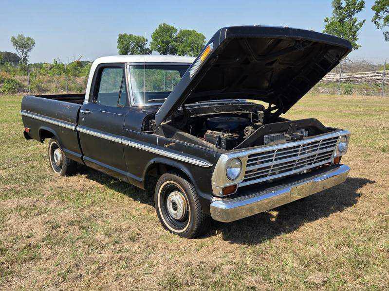 Black vintage pickup truck with its hood open, parked on dry grass in an outdoor setting with trees and blue sky in the background.