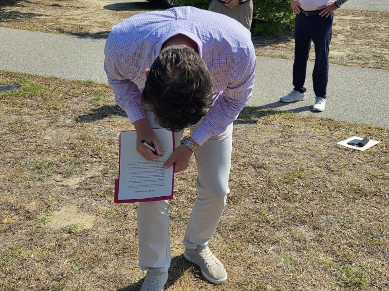 Man in a checkered shirt bends over, signing a document on a clipboard outdoors, with two other people standing in the background.