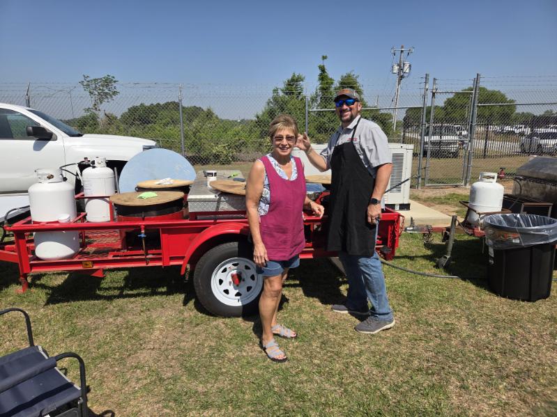Two people stand outdoors next to a red trailer with cooking equipment, both wearing aprons, smiling at the camera on a sunny day.