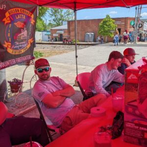 Several people sit under a red tent with a Dillon County Fire Department banner, at an outdoor event near a street and railroad tracks in a small town.