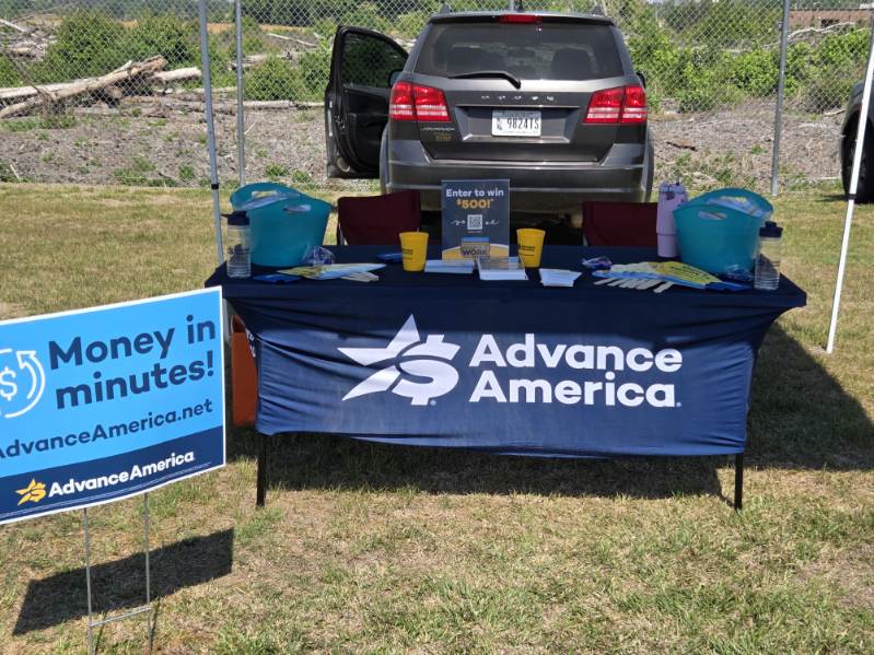 Advance America promotional table outdoors with informational materials, blue tablecloth, and sign reading "Money in Minutes!" in front of a parked SUV.