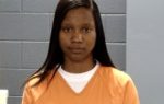 Woman with long straight hair wearing an orange prison uniform, standing against a gray cinderblock wall, looking directly at the camera.