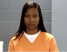Woman with long straight hair wearing an orange prison uniform, standing against a gray cinderblock wall, looking directly at the camera.