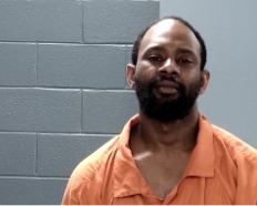 A man wearing an orange prison jumpsuit stands against a gray cinderblock wall, looking slightly upward with a neutral expression.