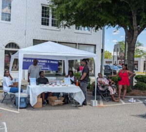 People gather around a white tented booth selling baked goods and drinks on a sunny day, with a banner reading "NYLA'S KREATIONZ & MORE" displayed.