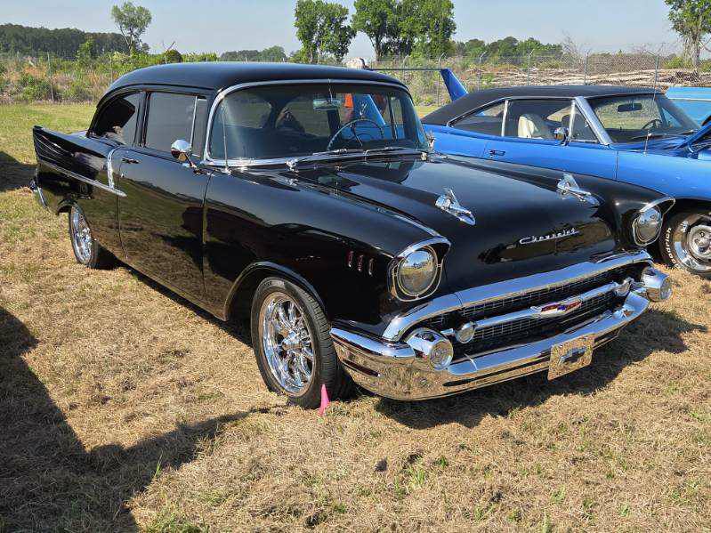 A shiny black vintage car parked on grass beside a blue classic car, with trees and other vehicles in the background on a sunny day.