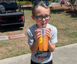 Young boy wearing glasses and a t-shirt drinks orange beverage from a plastic cup through a straw outdoors on a sunny day.