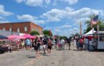 People walking around at an outdoor street fair with vendor tents, brick buildings, and a blue sky with scattered clouds.