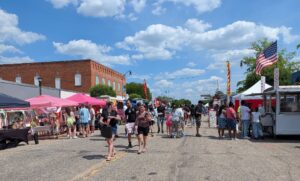 People stroll through a sunny outdoor street market with food trucks, vendor tents, and an American flag, under a blue sky with scattered clouds.