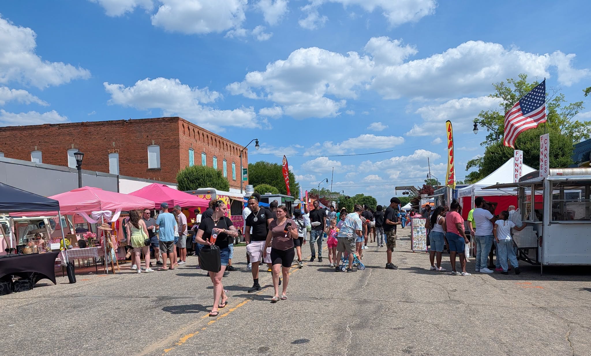People walking around at an outdoor street fair with vendor tents, brick buildings, and a blue sky with scattered clouds.