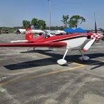 A small red and white aerobatic airplane is parked on an asphalt surface at an outdoor airfield under a clear blue sky.