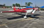 A small red and white aerobatic airplane is parked on an asphalt surface at an outdoor airfield under a clear blue sky.