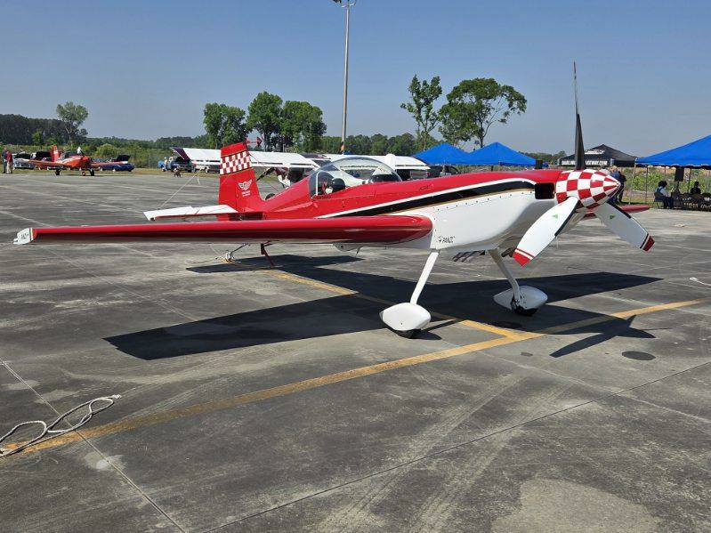 A small red and white aerobatic airplane is parked on an asphalt surface at an outdoor airfield under a clear blue sky.