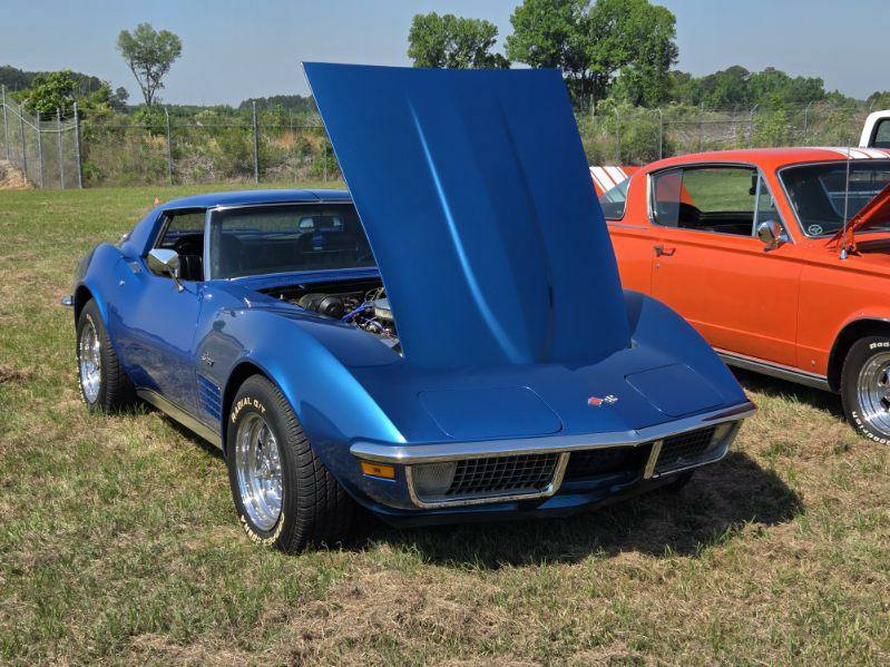 A blue classic car with its hood open, parked on grass next to an orange vintage car, under a clear sky.
