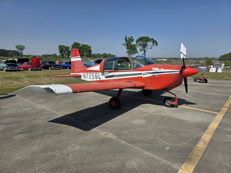 Red and white single-engine propeller airplane parked on a concrete surface, with trees and cars visible in the background under a clear blue sky.