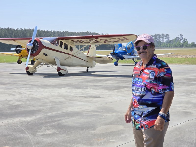 A man in a colorful shirt and cap stands on an airport runway with vintage airplanes in the background on a sunny day.