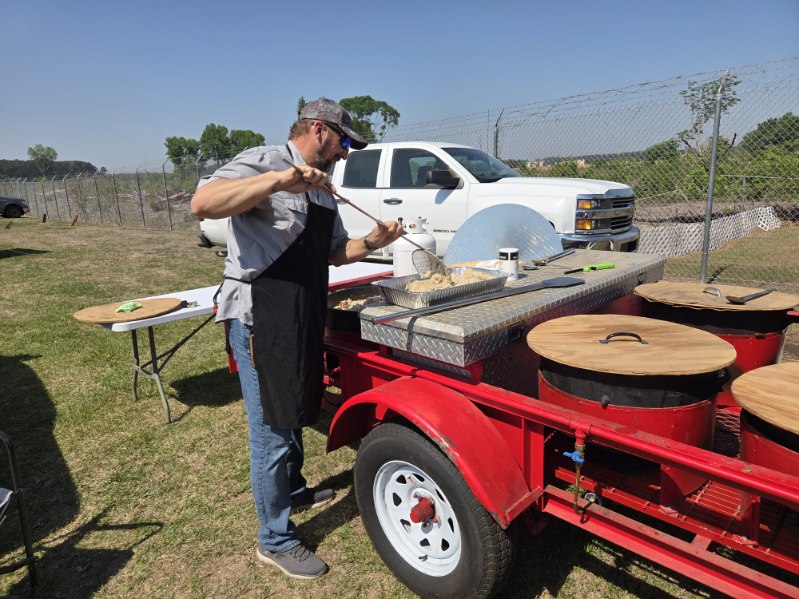 A man in an apron cooks outdoors beside a red grill trailer, preparing food outdoors with a white pickup truck parked nearby.