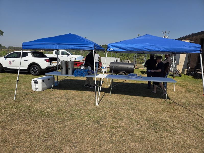 Two blue canopy tents set up outdoors on grass, with tables underneath and people standing behind, next to parked white vehicles on a sunny day.