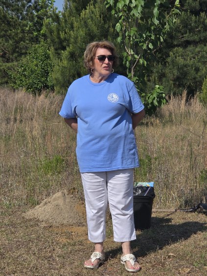 Woman wearing sunglasses, a blue T-shirt, and white pants stands outdoors on a grassy area with trees and a small mound of dirt behind her.