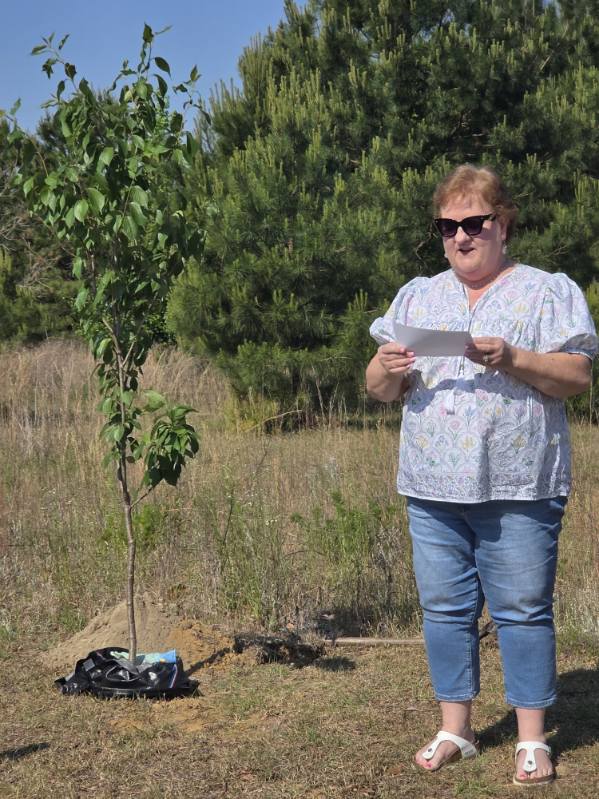A woman stands outdoors near a newly planted tree, reading from a paper. She is wearing sunglasses, a patterned blouse, and jeans. Pine trees are in the background.