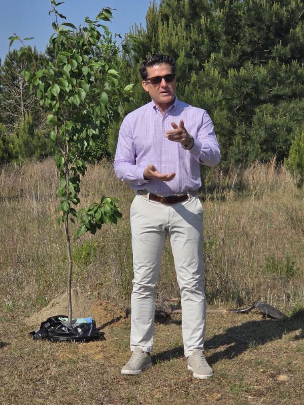 A man in sunglasses and light clothes speaks outdoors near a small tree on a grassy field with forest in the background.