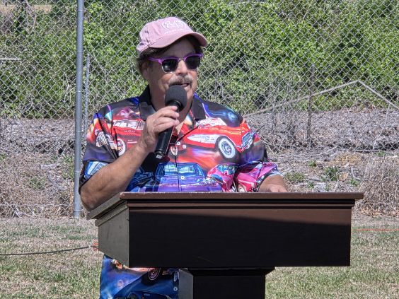 Man in colorful shirt and pink cap speaking into a microphone at an outdoor podium, with greenery and a fence in the background.