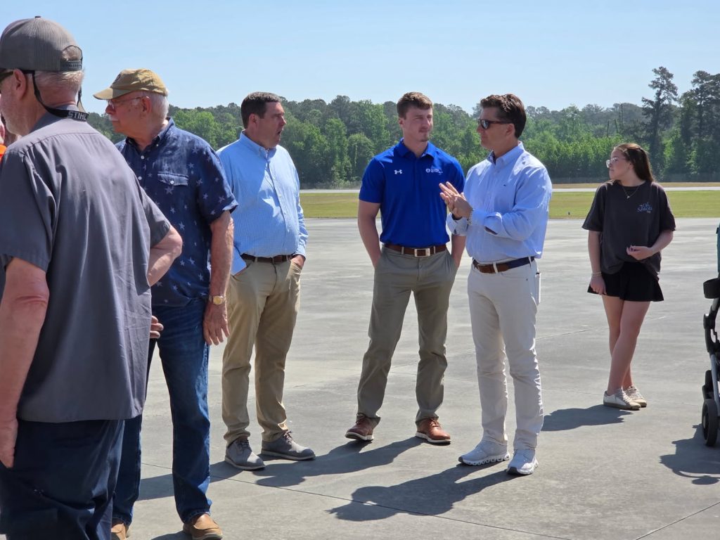 A group of people standing and talking outdoors on a sunny day, with trees and an open area in the background.